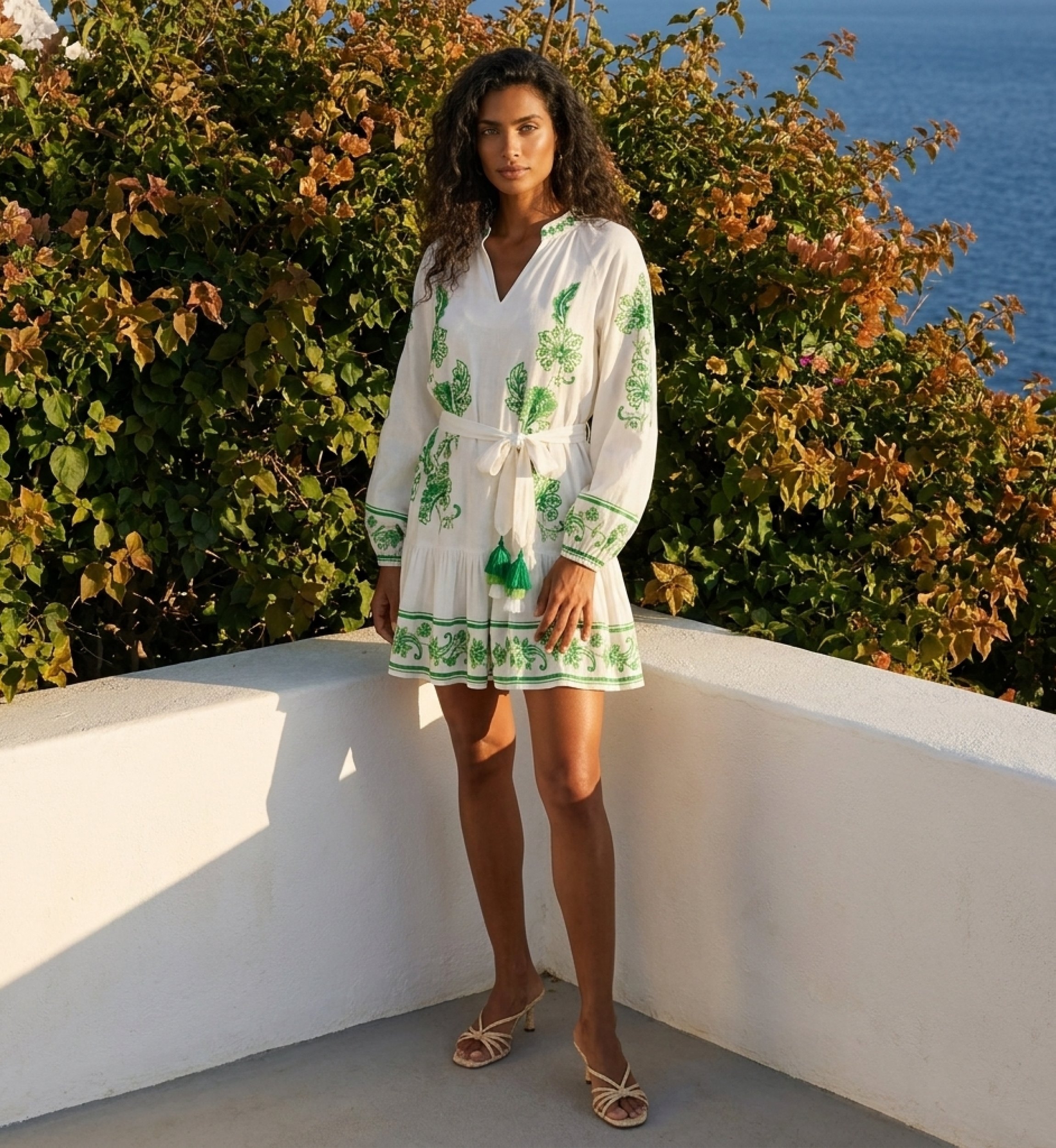 Vestido curto branco manga comprida com bordado floral verde, decote em V, cintura amarrada com borlas e saia em camadas.