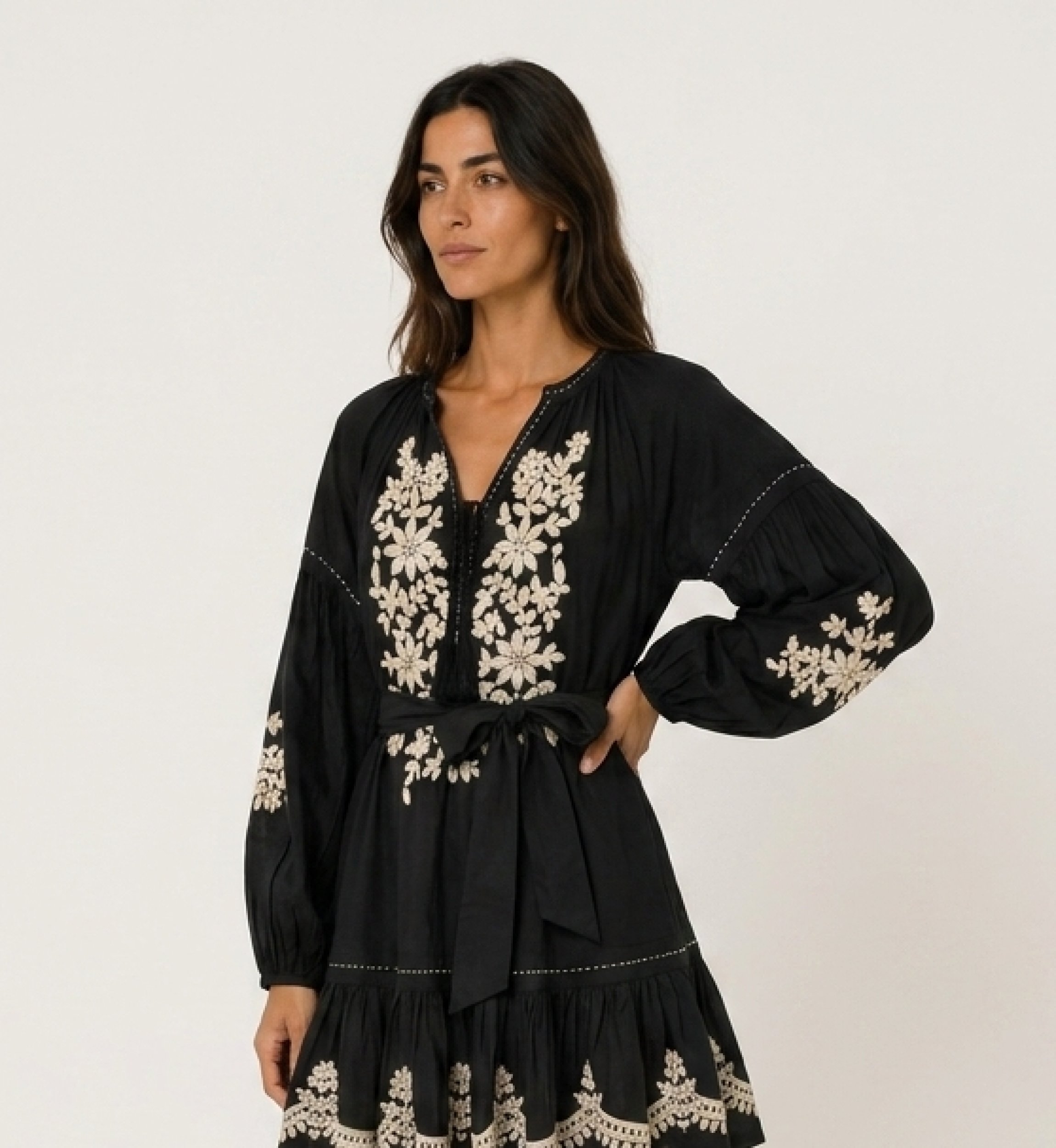 Vestido mini negro holgado con mangas largas abullonadas, falda escalonada, bordado floral blanco y cintura anudada.