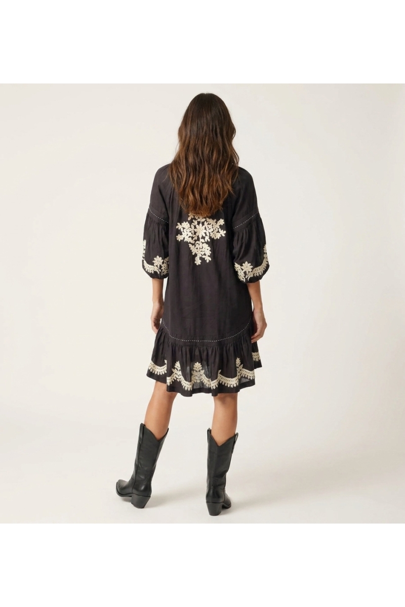 Vestido mini preto solto, mangas balão longas, saia em camadas, bordado floral branco, cintura com laço, vista traseira.