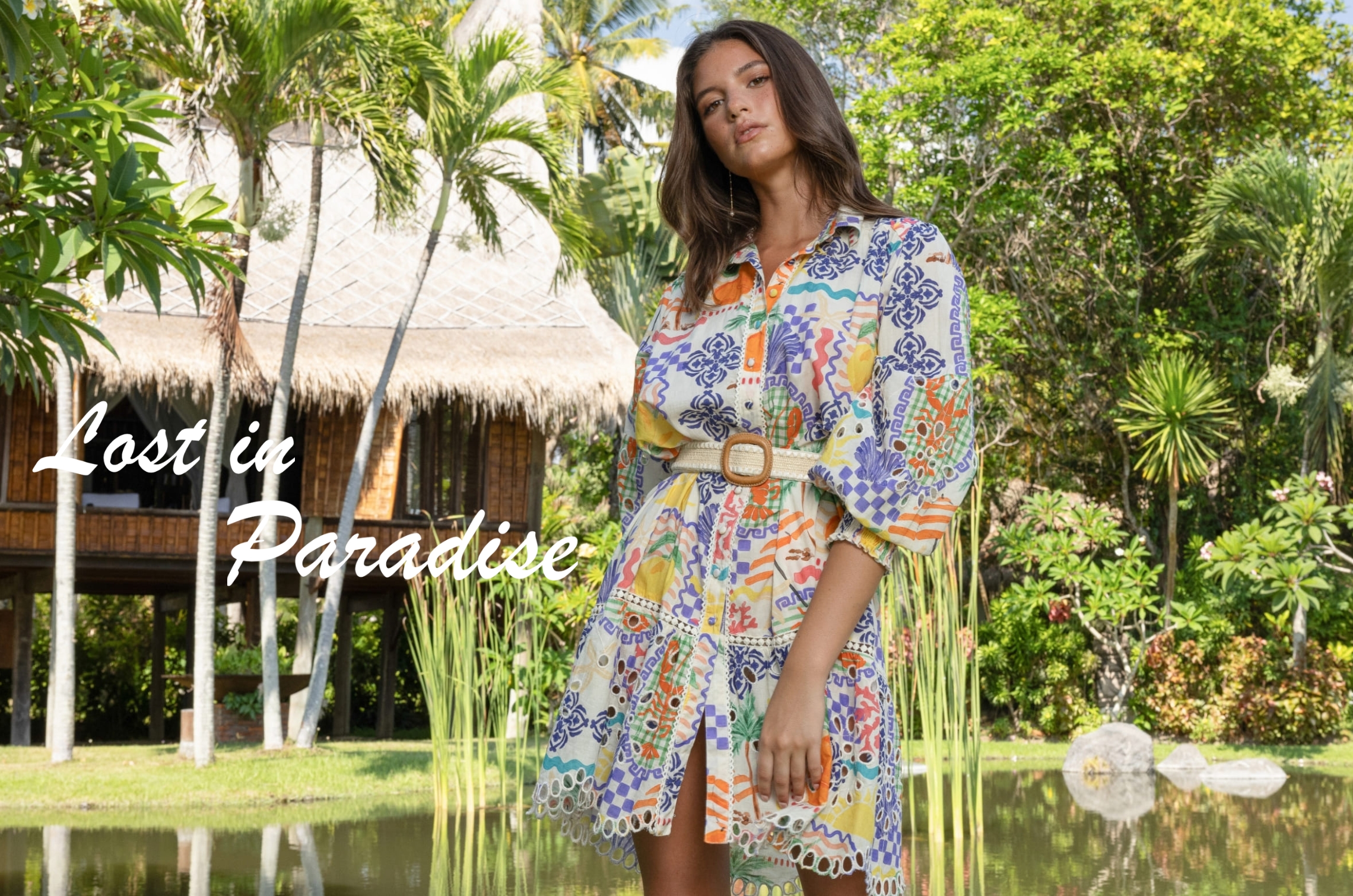 Mannequin portant une robe chemise courte multicolore ceinturée, dans un jardin tropical devant un bungalow, avec le texte « Lost in Paradise ».