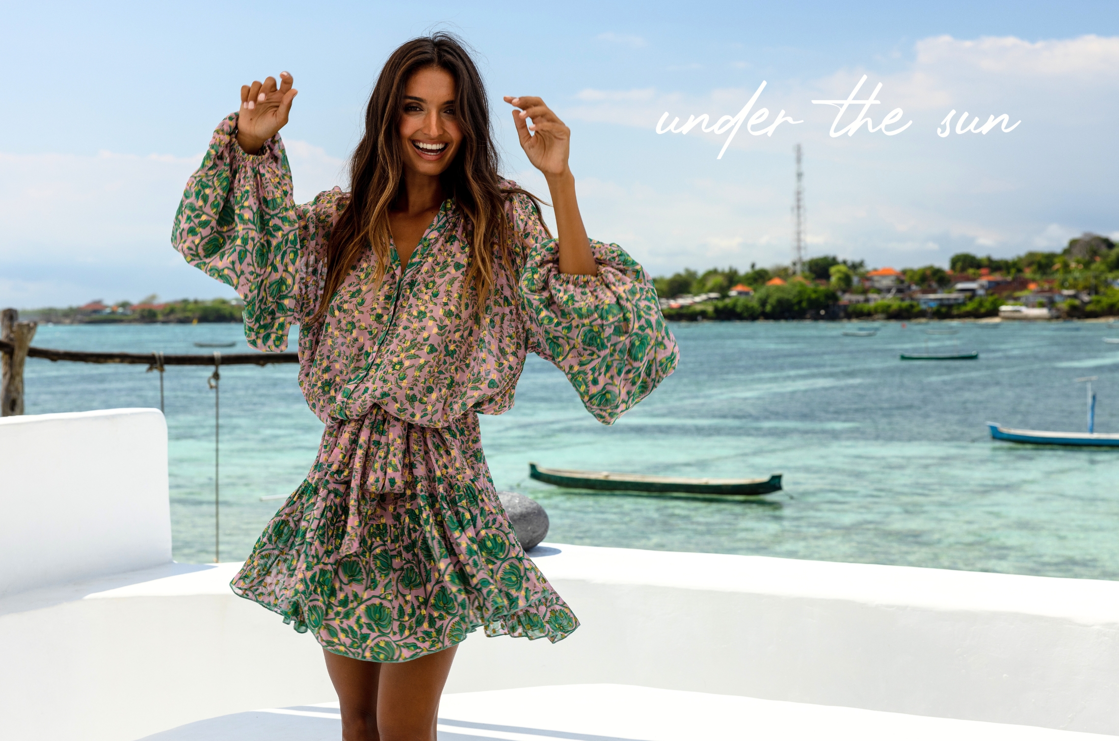 Mannequin souriant portant une robe courte rose à imprimé vert, sur une terrasse face à la mer turquoise avec des bateaux, avec le texte « under the sun ».
