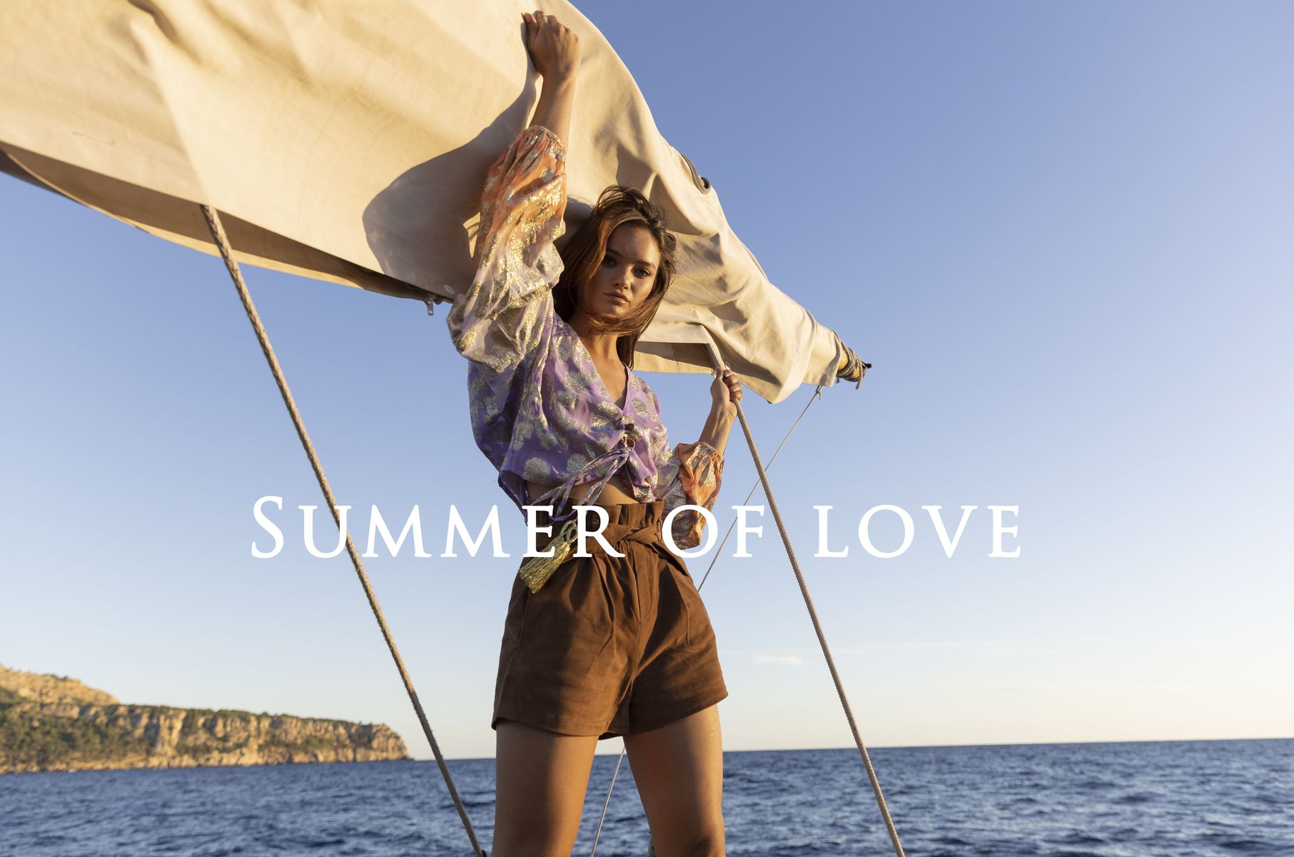 Mannequin portant une blouse violette imprimée et un short marron sur un voilier en bord de mer, avec le texte « SUMMER OF LOVE ».