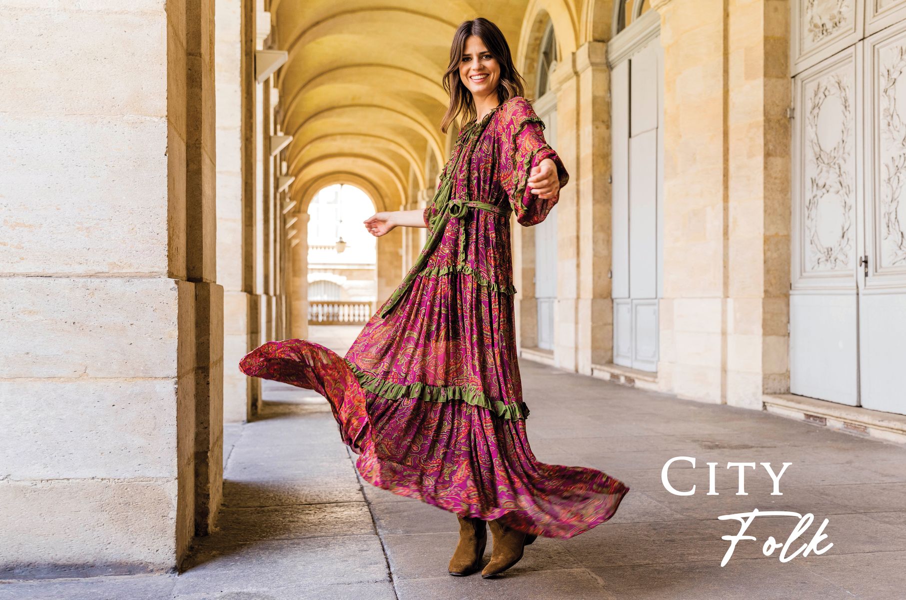 Mannequin souriant portant une robe longue bohème fuchsia et verte, tournoyant sous des arcades en pierre, avec le texte « City Folk ».