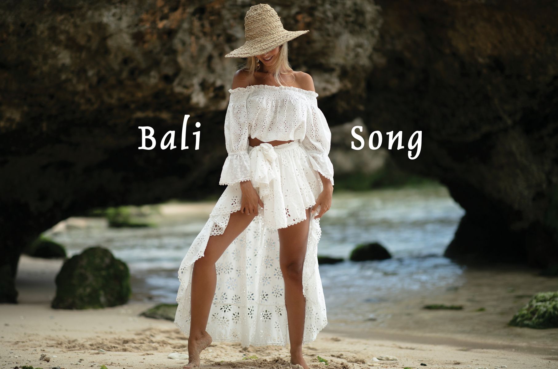 Mannequin portant un ensemble blanc en broderie anglaise avec top épaules dénudées et jupe longue fendue, sur une plage rocheuse, avec le texte « Bali Song ».