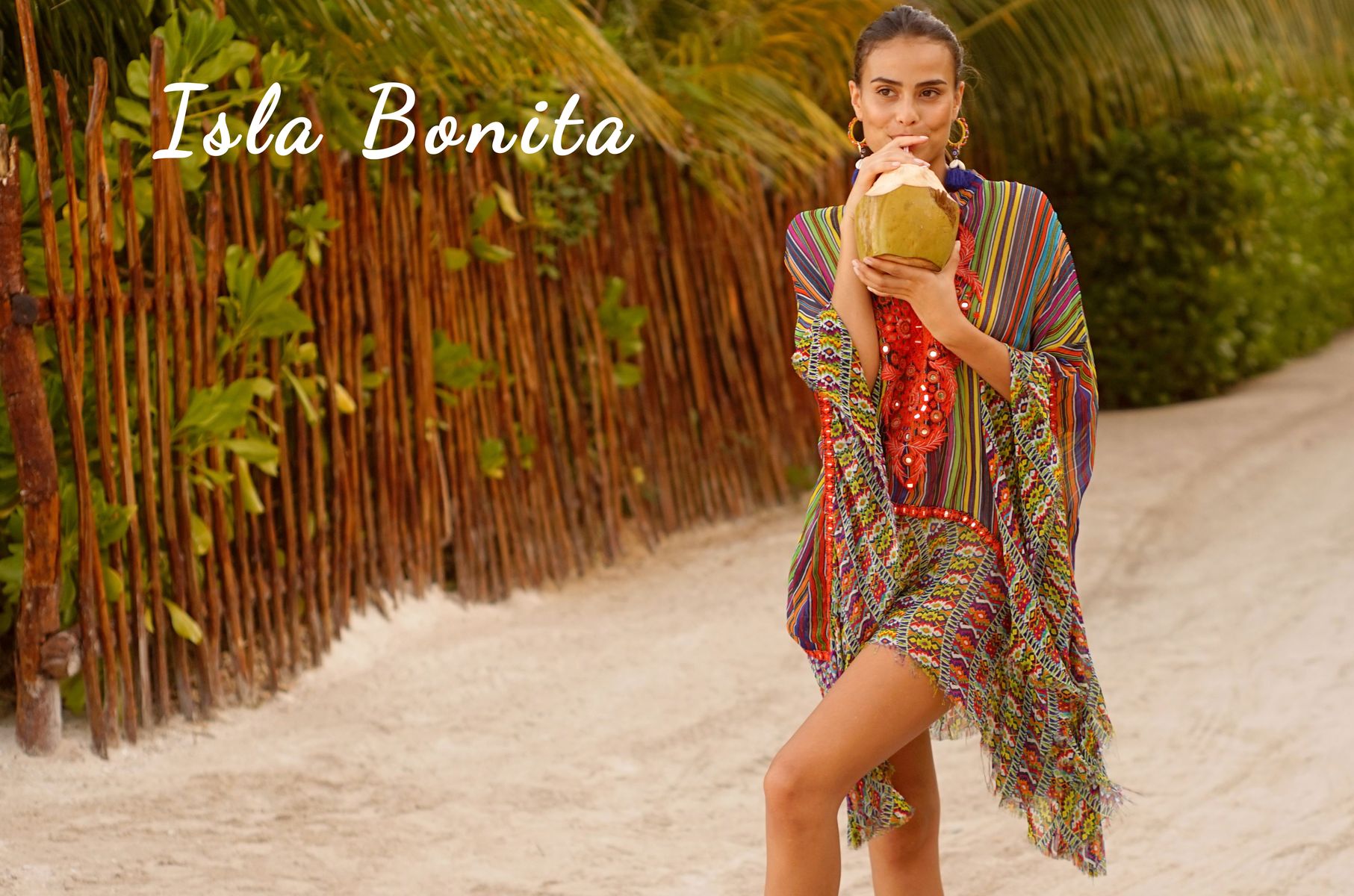 Mannequin portant un kaftan multicolore à broderies rouges, marchant sur un chemin de sable en tenant une noix de coco, avec le texte « Isla Bonita ».