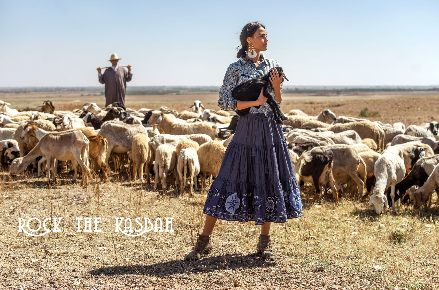 Mannequin portant une blouse imprimée et une jupe longue bleue, tenant un agneau devant un troupeau dans un paysage aride, avec le texte « Rock the Kasbah ».