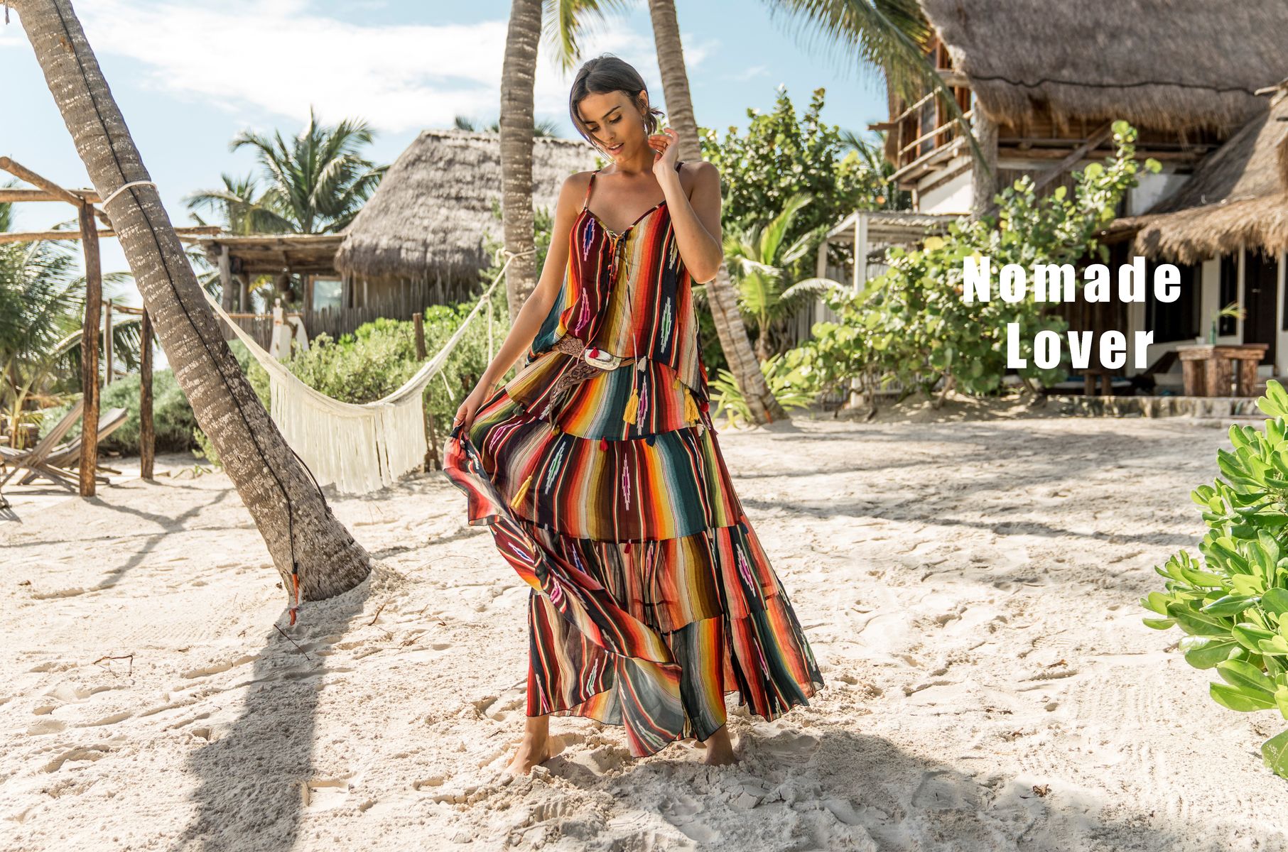 Mannequin portant une robe longue à rayures multicolores sur une plage avec palmiers et cabanes, avec le texte « Nomade Lover ».